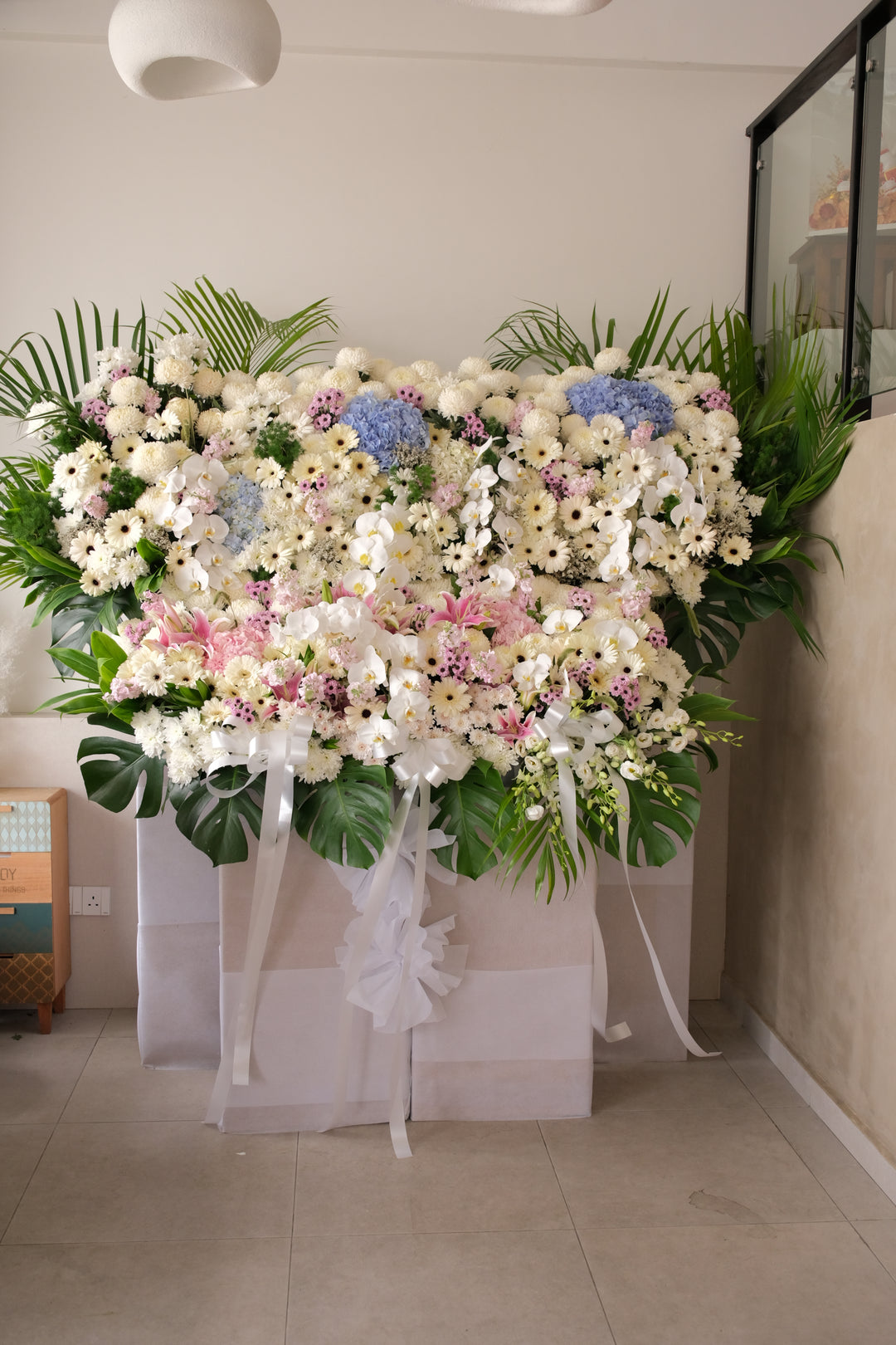 A large condolence flower arrangement styled like a garden, featuring pastel blooms and lush layers. Displayed as a standing piece, designed to honour a life and offer a soft farewell.