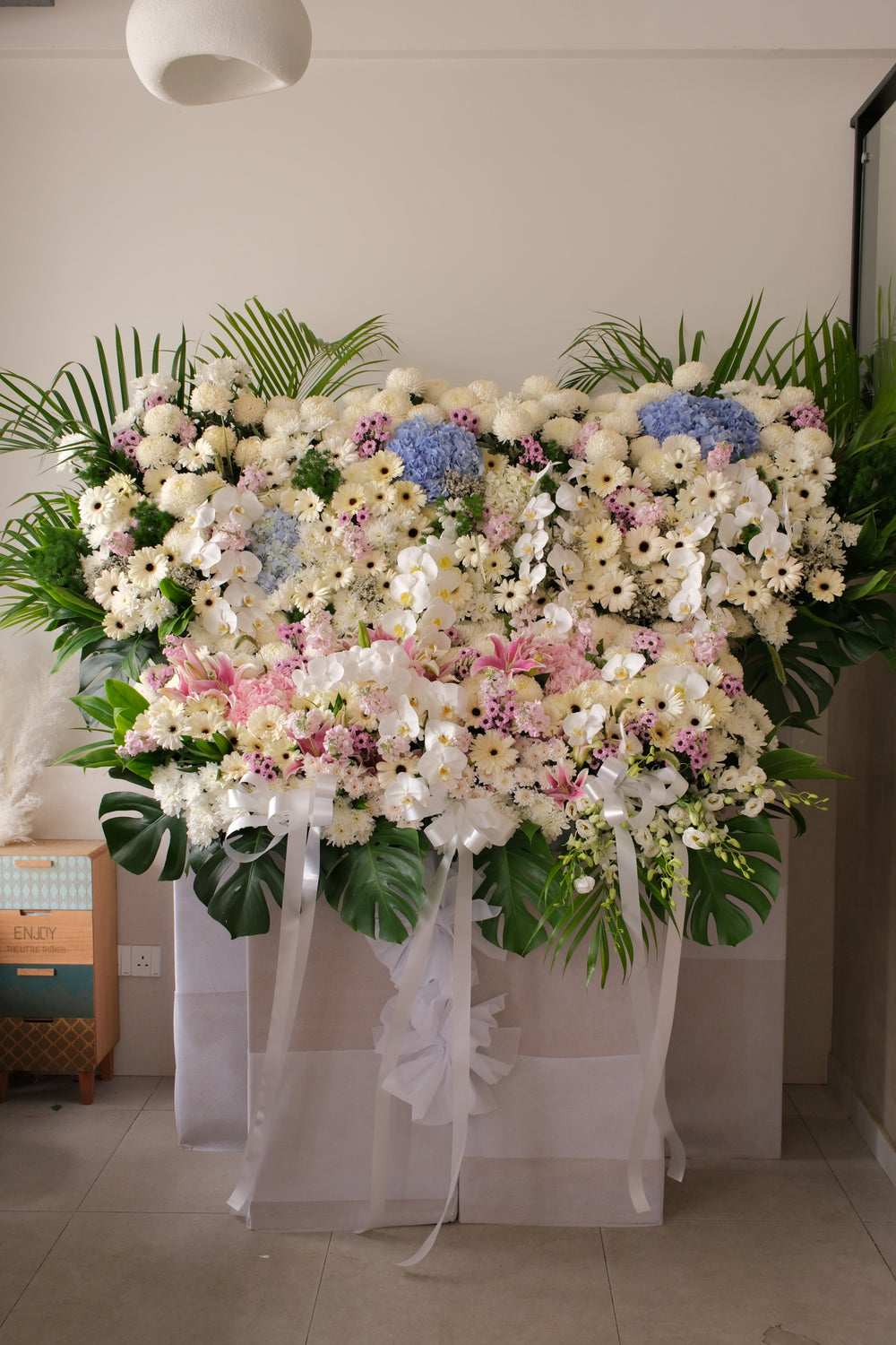 A large condolence flower arrangement styled like a garden, featuring pastel blooms and lush layers. Displayed as a standing piece, designed to honour a life and offer a soft farewell.