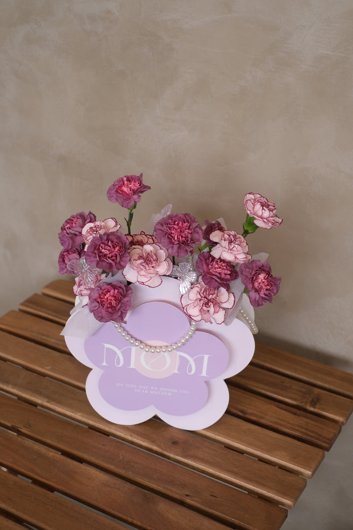 A luxurious box filled with 16 stalks of vibrant purple carnations, arranged beautifully and adorned with shimmering pearls. The flowers exude elegance and grace, symbolizing admiration and gratitude for motherhood. The box is set against a backdrop of soft lighting, accentuating its beauty. Text overlay reads 'Matriarchal Magnificence: 16 Stalks of Purple Carnations in a Pearl-Adorned Box.' Delivery available for Mother's Day in Penang
