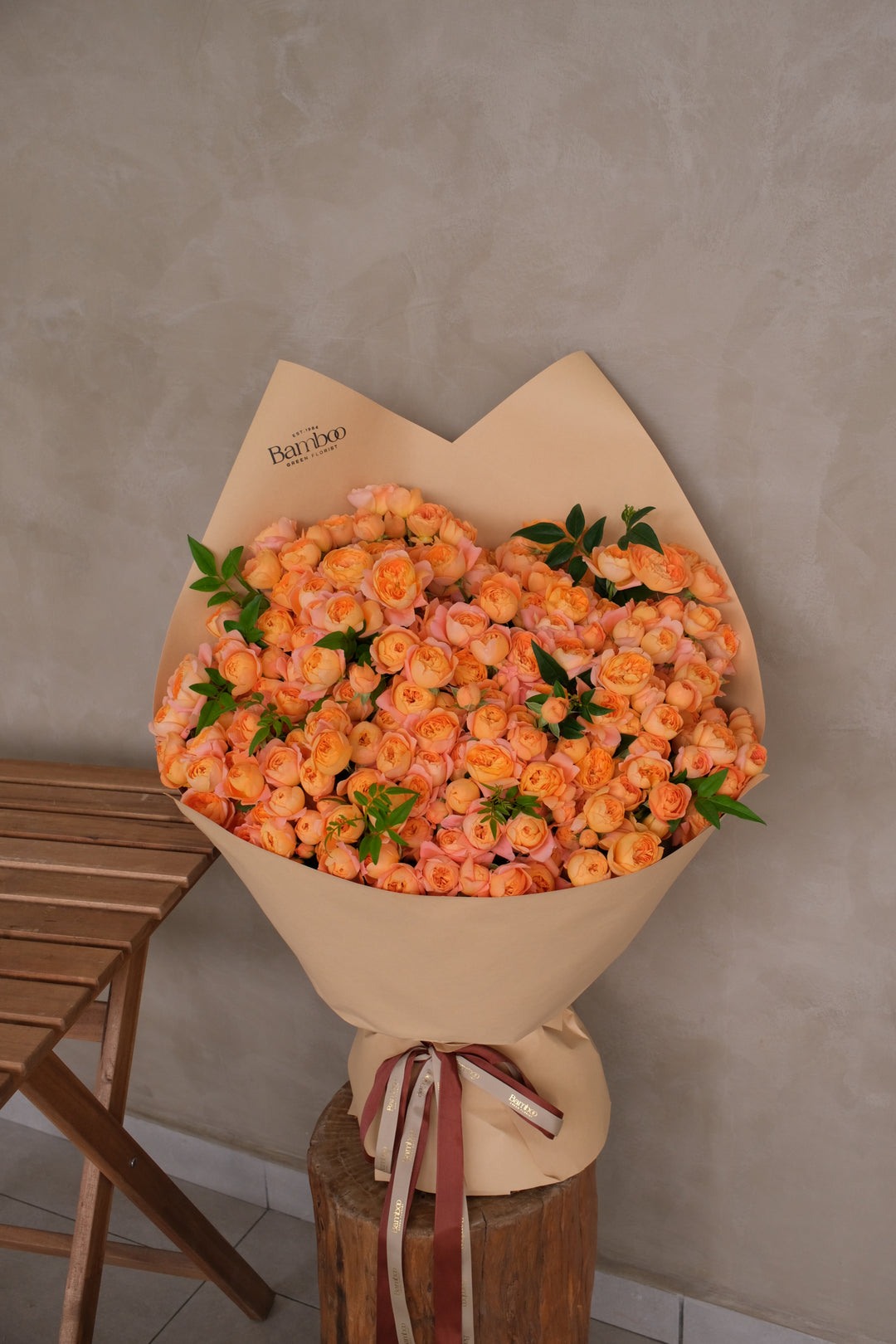 A minimal bouquet of coral-toned multi-head garden roses, paired only with green leaves and wrapped in kraft paper. Natural and luxe in appearance.