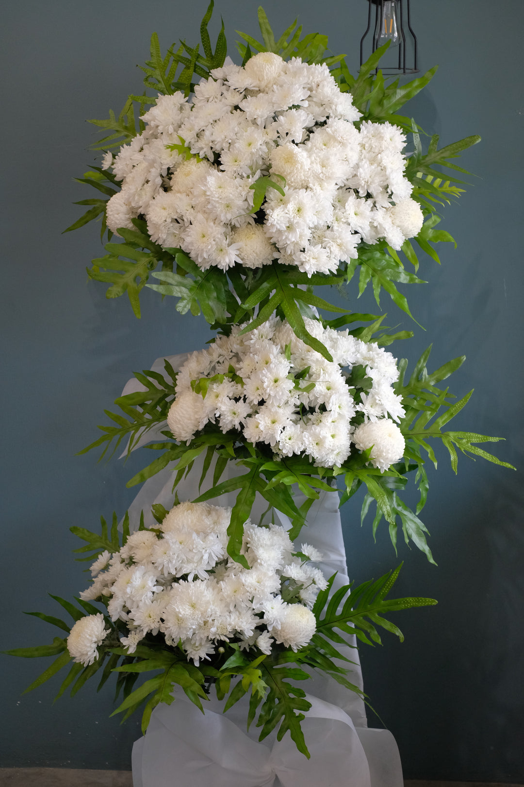 3 Tier standing wreath with all white poms and greens. Decorate in purple and white in honor and remembrance of the dearly departed. For same day condolences flowers delivery in Penang.