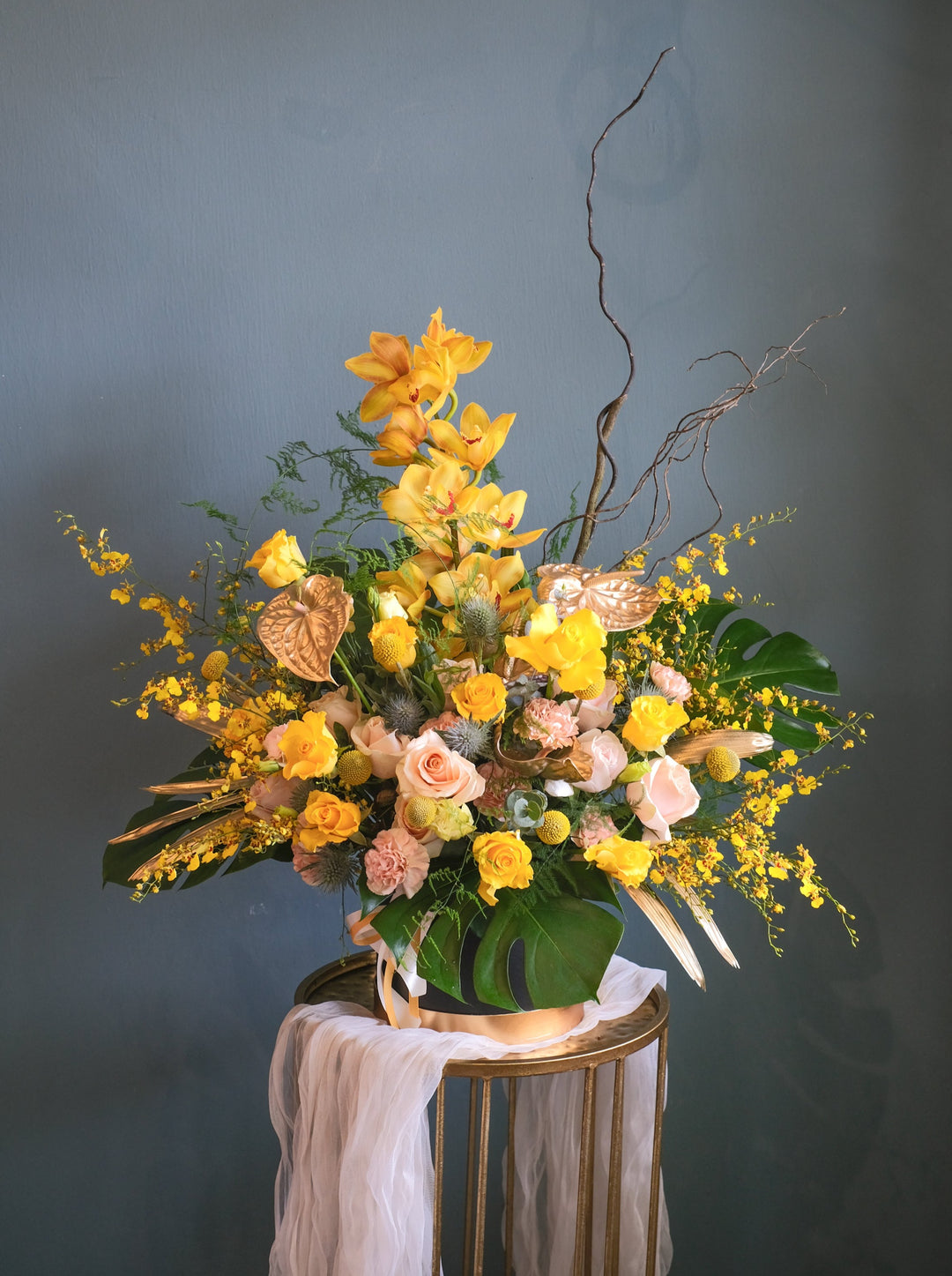 A gorgeous vibrant flower arrangement in a Golden Paradise theme, featuring Cymbidium orchids, golden yellow roses, carnations, yellow craspedia, eryngium, and lush leaves.