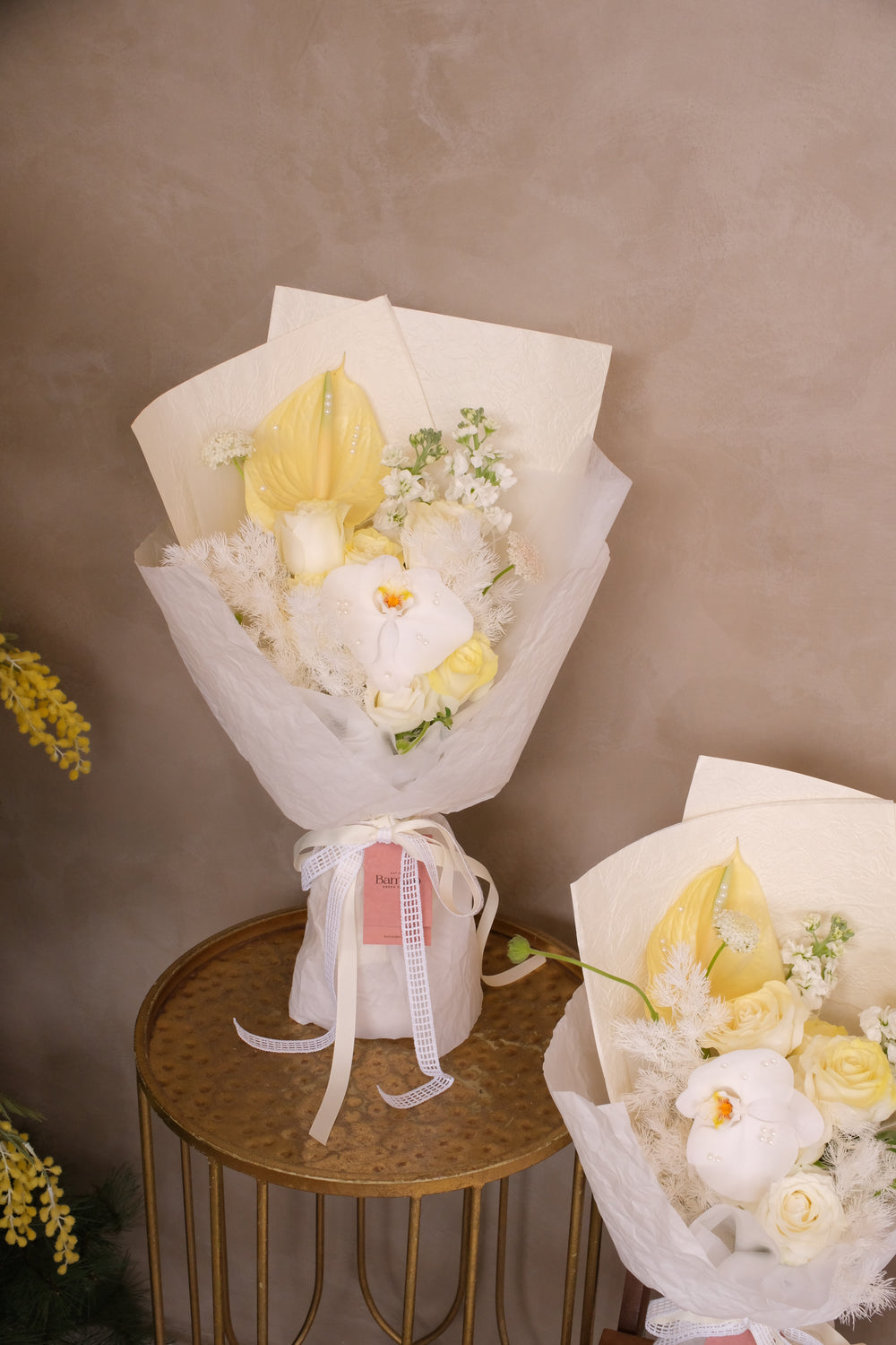 Cream and pale yellow bouquet with white orchid, anthurium and roses, wrapped in white crushed paper on a brass side table.