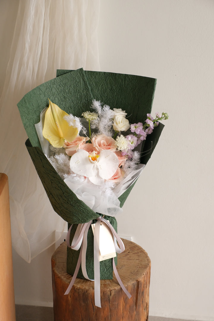 Pastel bouquet with white orchid and pearls, wrapped in dark green crushed paper, standing on a wooden stump.