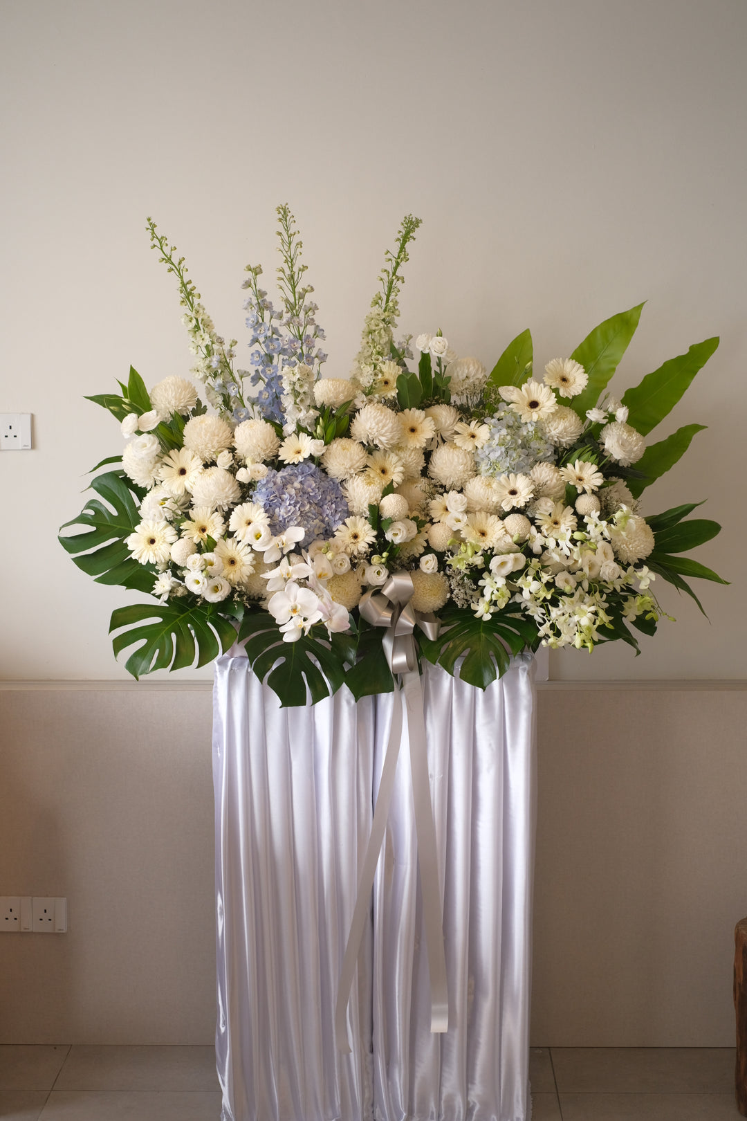 A serene arrangement of white roses, pink carnations, and blue forget-me-nots, symbolizing peace, remembrance, and eternal love, placed in a tasteful stand for penang delivery