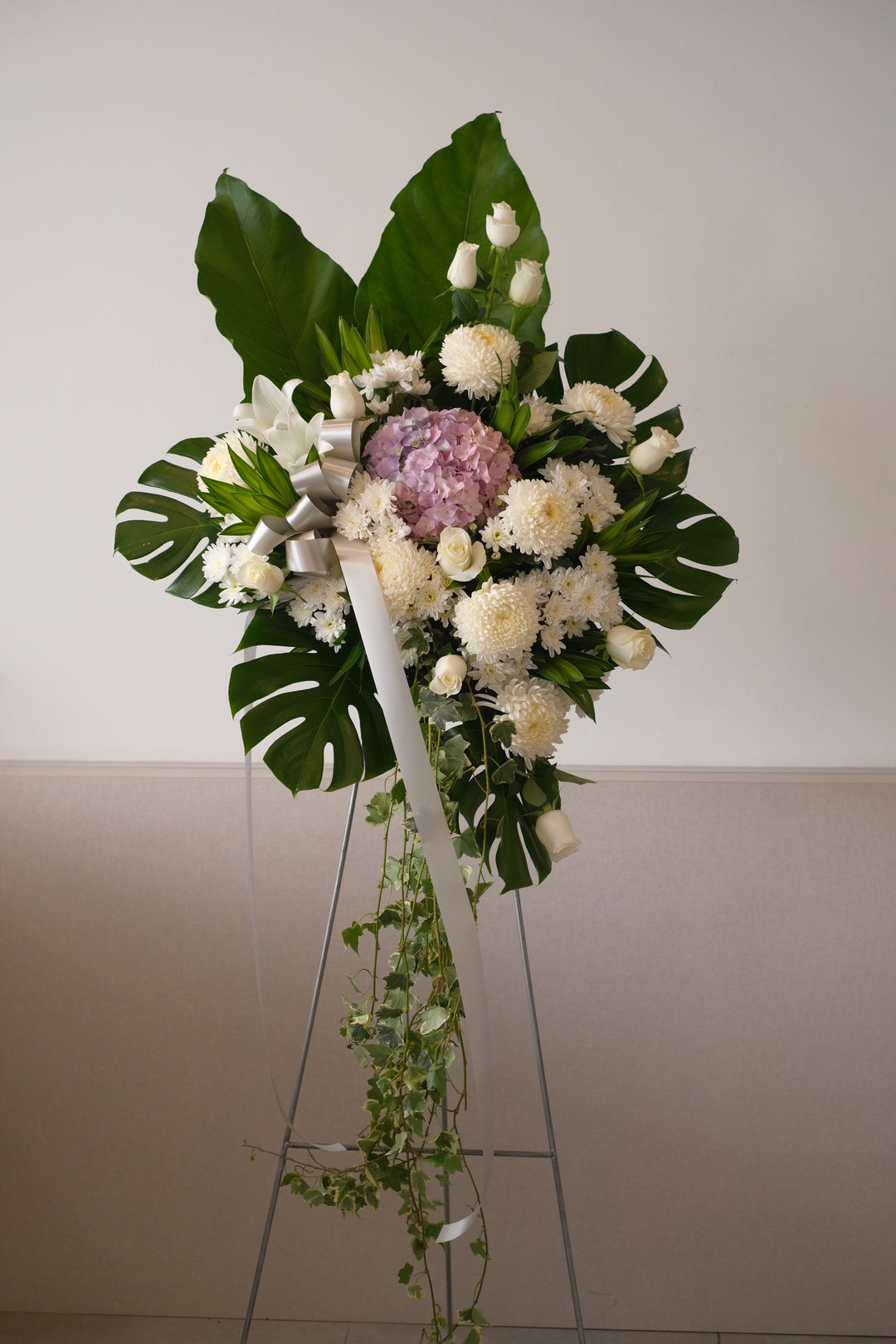 Pure, white and ethereal condolence stand featuring cream roses, white chrysanthemum and hydrangea. For same day condolences flowers delivery in Penang.  