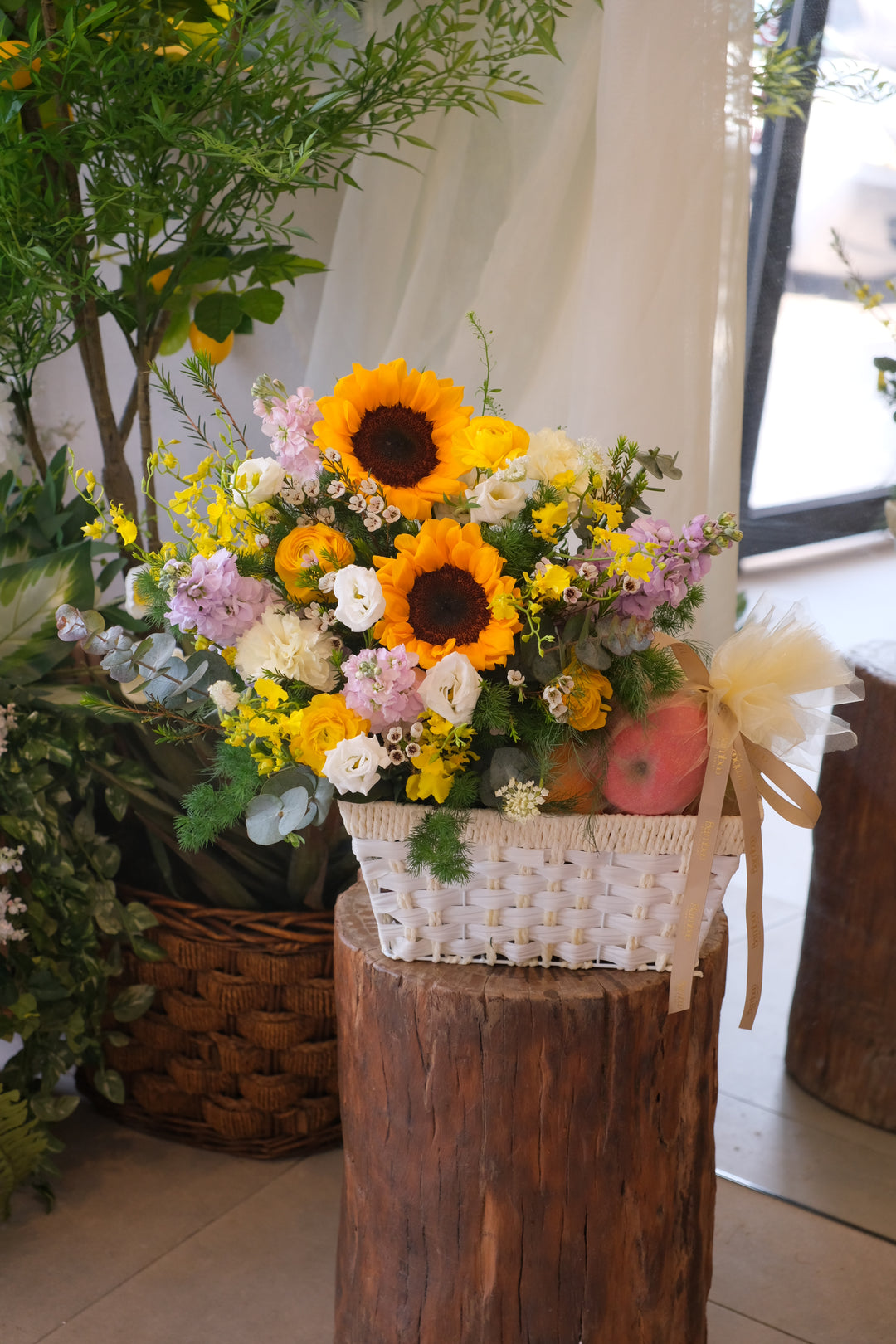 Rustic white wicker basket filled with sunflowers, purple stock flowers, and fresh apples on a wooden stump, by Bamboo Green Florist.