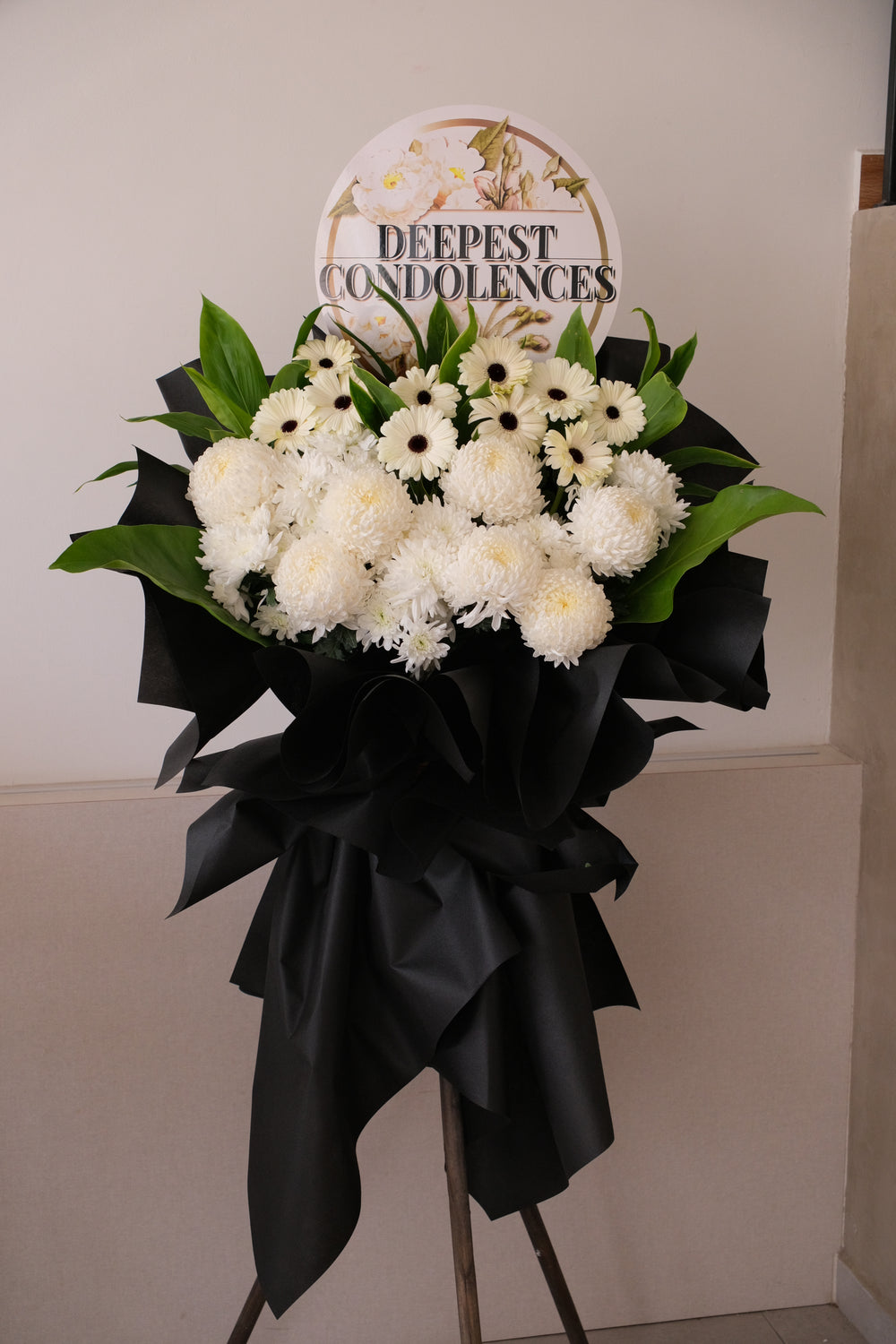 Gentle Reflections condolence floral stand with white ping pong chrysanthemums and white gerberas in professional black wrapping on a tripod.