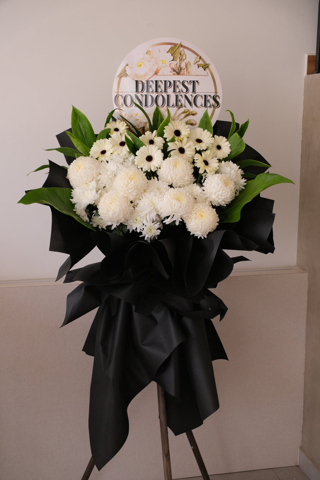 Gentle Reflections condolence floral stand with white ping pong chrysanthemums and white gerberas in professional black wrapping on a tripod.