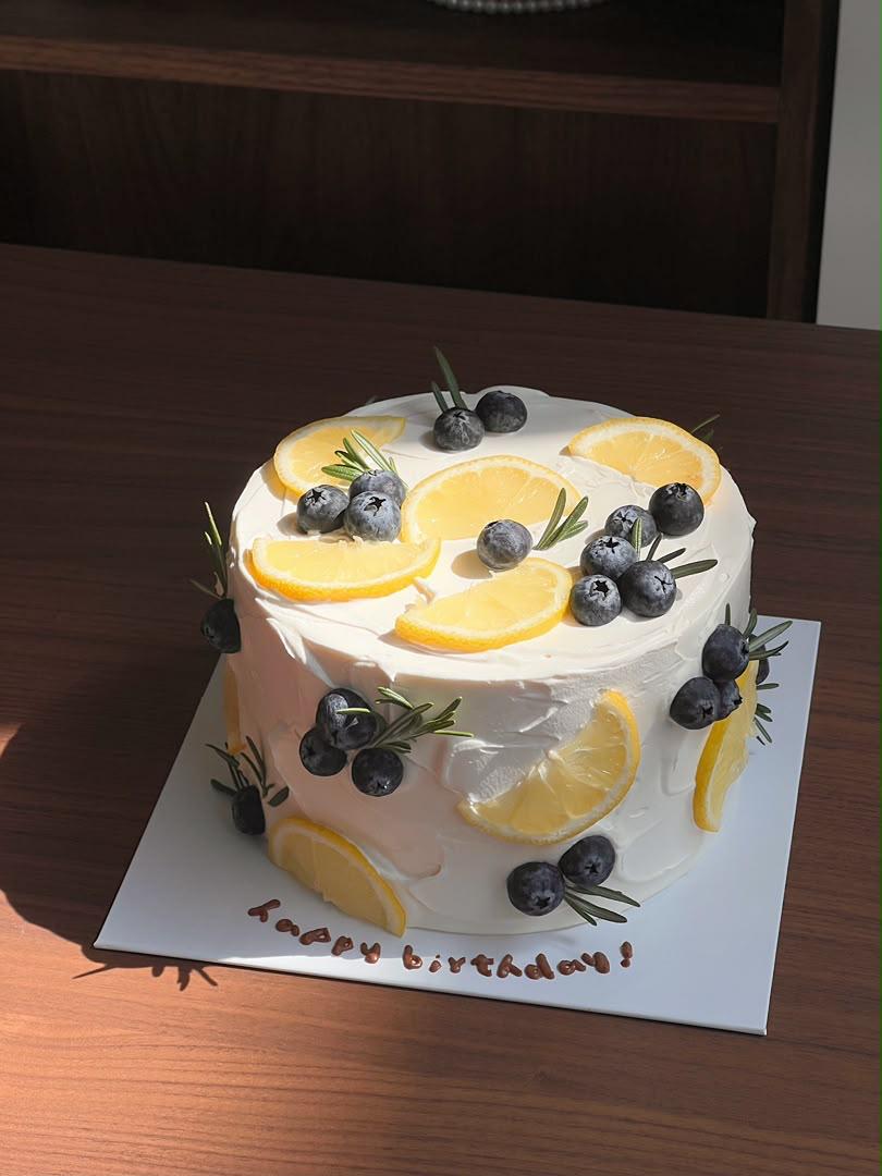 Lemon cream cheese cake with white frosting, topped with fresh lemon slices, blueberries and rosemary sprigs, on a white board over a wooden table.
