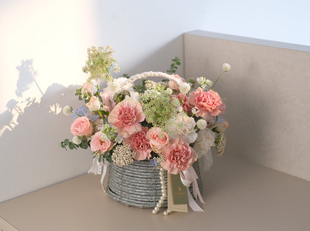 A beautiful collection of springtime flowers, including carnations, pink rose spray, mini delphiniums, rice flowers,  arranged in a rustic wooden basket.
