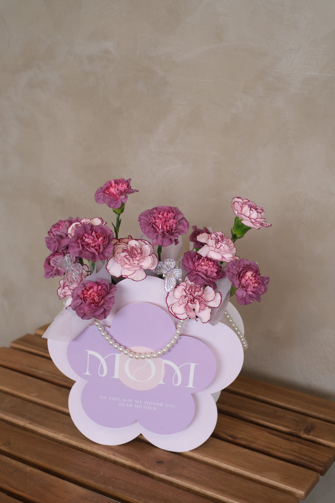 A luxurious box filled with 16 stalks of vibrant purple carnations, arranged beautifully and adorned with shimmering pearls. The flowers exude elegance and grace, symbolizing admiration and gratitude for motherhood. The box is set against a backdrop of soft lighting, accentuating its beauty. Text overlay reads 'Matriarchal Magnificence: 16 Stalks of Purple Carnations in a Pearl-Adorned Box.' Delivery available for Mother's Day in Penang