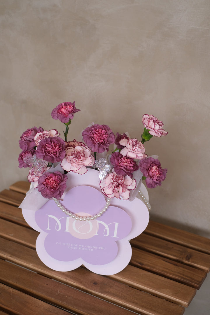 A luxurious box filled with 16 stalks of vibrant purple carnations, arranged beautifully and adorned with shimmering pearls. The flowers exude elegance and grace, symbolizing admiration and gratitude for motherhood. The box is set against a backdrop of soft lighting, accentuating its beauty. Text overlay reads 'Matriarchal Magnificence: 16 Stalks of Purple Carnations in a Pearl-Adorned Box.' Delivery available for Mother's Day in Penang