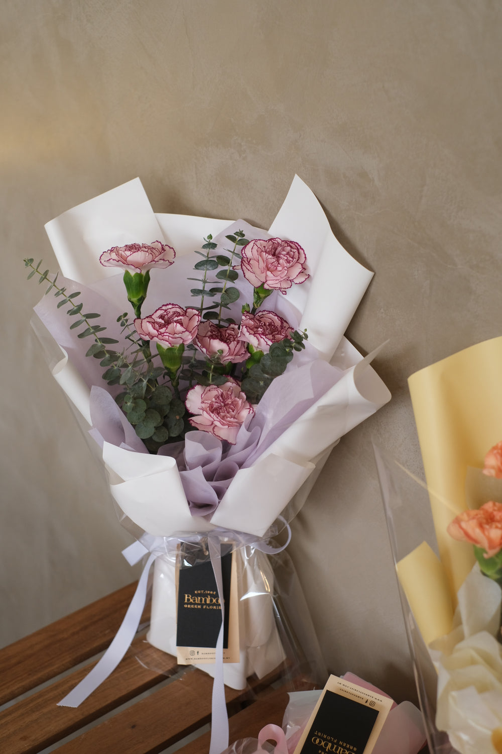 A breathtaking floral arrangement titled 'Stardust Carnation Ensemble' featuring a mesmerizing blend of orange, purple, and pink carnations, arranged elegantly in a vase. Each bloom shines like stardust, radiating warmth and enchantment. Text overlay reads 'Stardust Carnation Ensemble' by Bamboo Green Florist, your trusted Penang florist for exquisite Mother's Day blooms