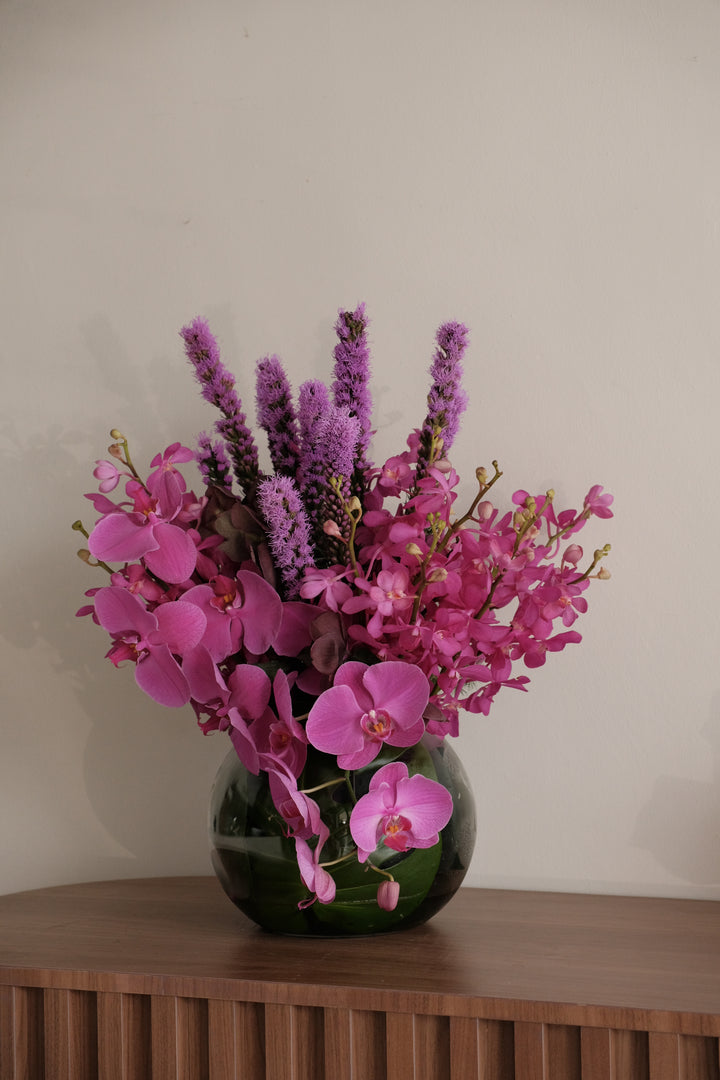 Modern monochromatic purple orchid arrangement with Phalaenopsis and Liatris in a round glass fishbowl vase.