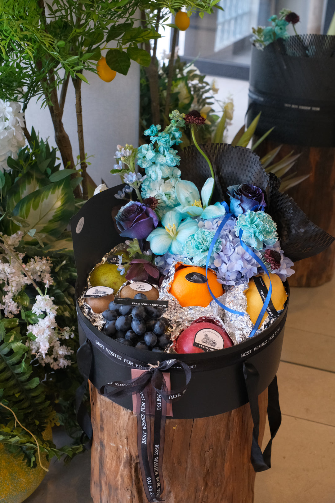 Luxury black gift box filled with fresh grapes, oranges, and apples paired with blue hydrangeas and roses by Bamboo Green Florist.