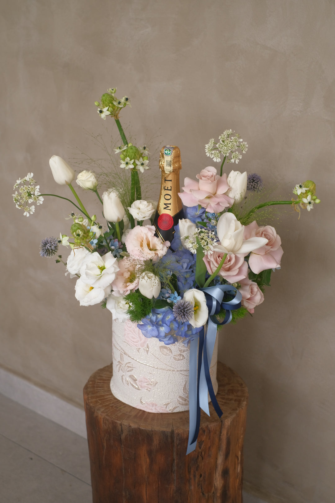 Elegant bloom box with pastel nude roses, blue-hued flowers, tulips, and a bottle of champagne Moet Chandon, perfect for special occasions like birthdays or anniversaries.