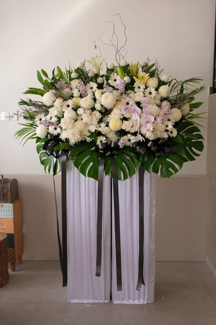 Side view of a condolence floral display in soft white and lavender tones, arranged with gentle height and foliage accents.