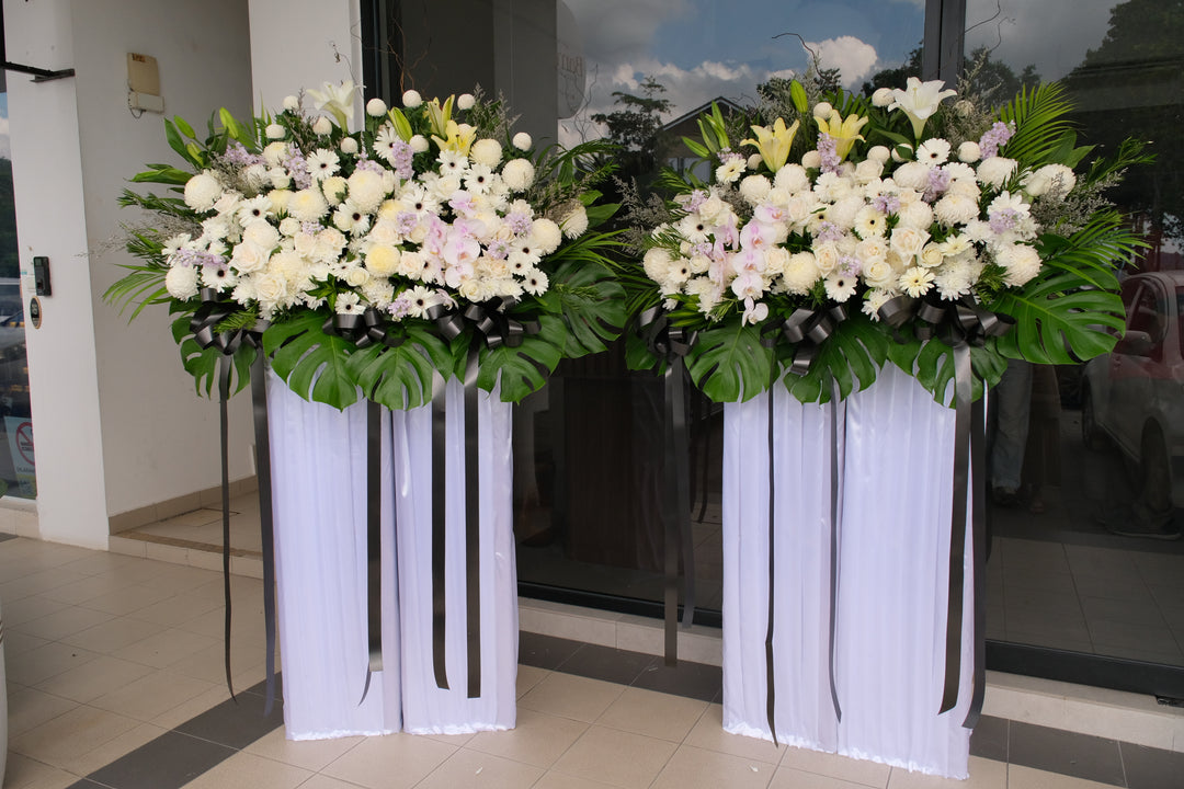 A condolence flower arrangement featuring white and soft lilac roses, matthiolas, daisies, and chrysanthemums, mounted on a structured floral box with decorative foliage.