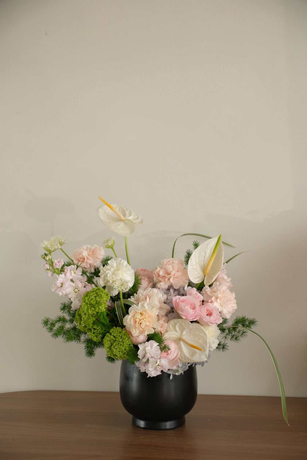Modern Mother's Day floral arrangement with white Anthuriums, blush pink roses, and carnations in a round black ceramic vase.