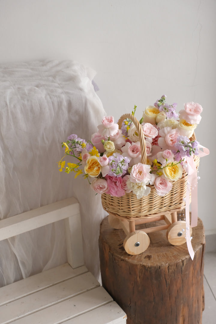 Tiny Treasures new baby flower basket in soft blush, cream, lavender and yellow garden blooms, arranged in a woven pram basket on wooden wheels.

