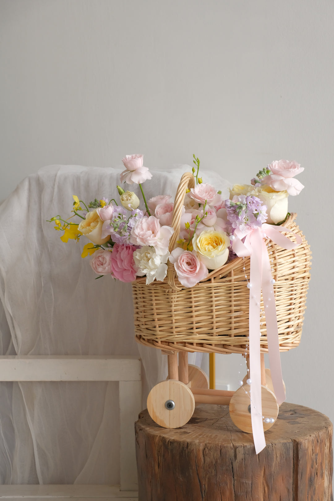 Side view of the First Light pram flower basket showing the wooden wheels, blush ribbon and trailing pearl detail.
