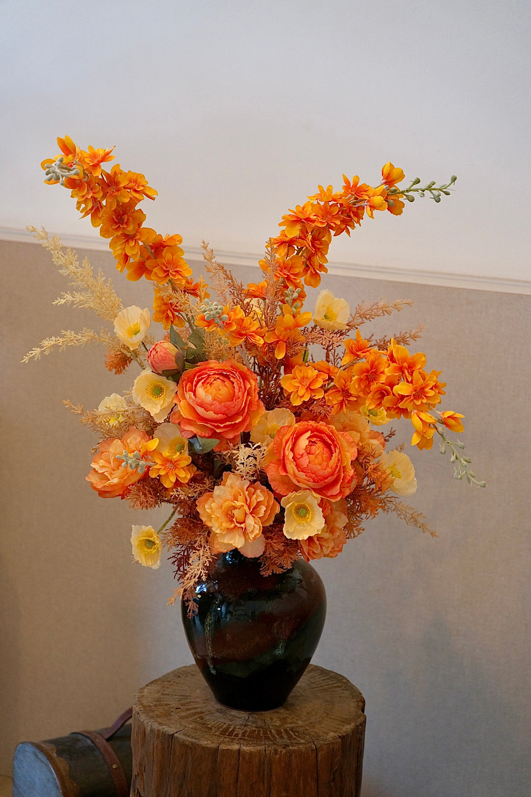 Artificial orange and apricot floral arrangement in a glossy ceramic vase, featuring lifelike ranunculus, poppies, and wild sprays on a tabletop.