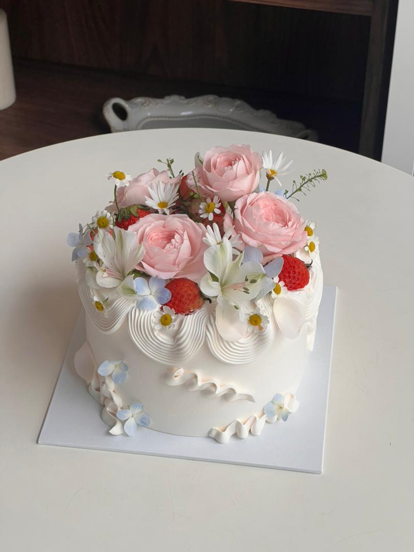 Flower Flora customized cake with fresh pink roses, white daisies, and strawberries on a white whipped cream base with scalloped piping.