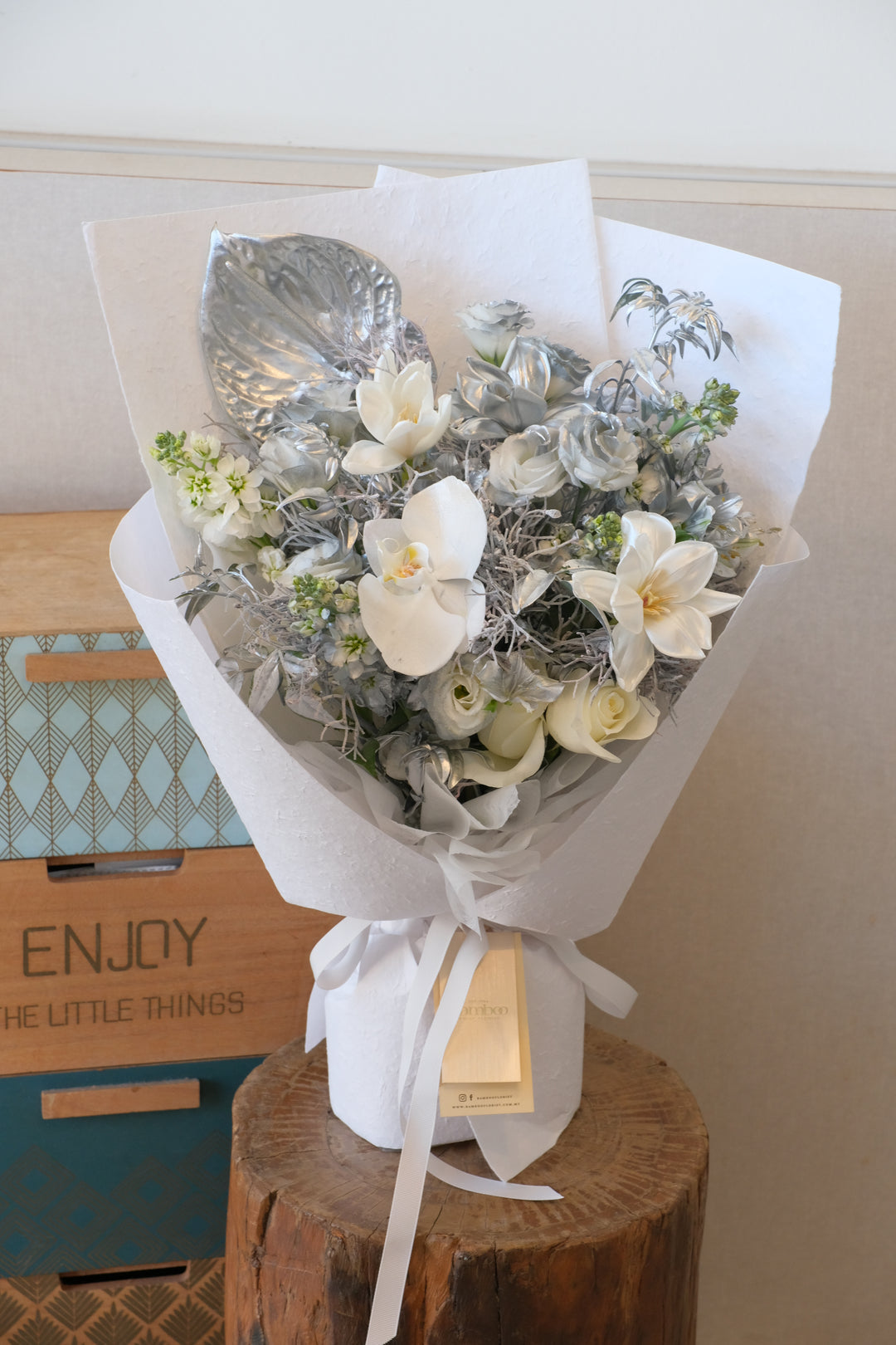Premium white Phalaenopsis orchid bouquet with metallic silver painted leaves and white roses in a minimalist white textured wrap.