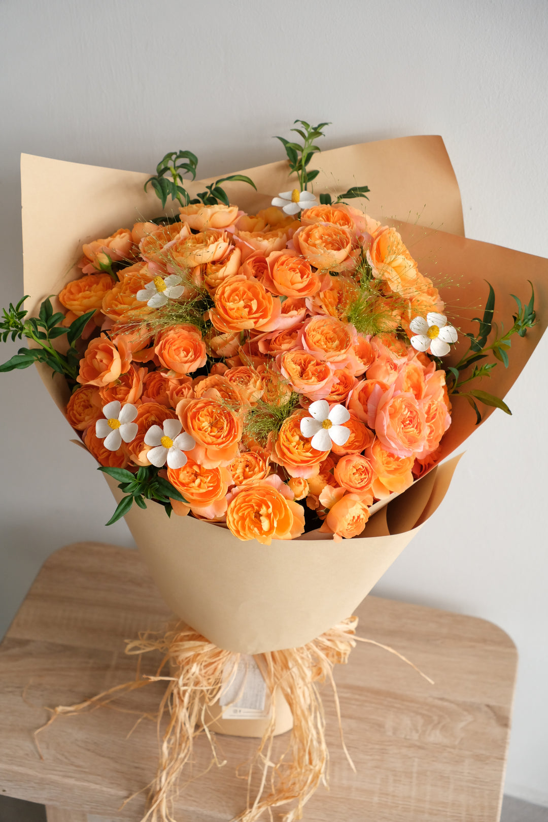 Coral-orange garden rose bouquet with white daisies and green grasses in kraft paper, hand-tied by Bamboo Green Florist Penang