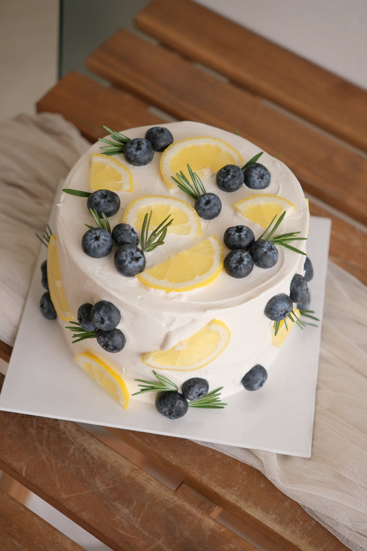 Lemon cream cheese cake with white frosting, topped with fresh lemon slices, blueberries and rosemary sprigs, on a white board over a wooden table.