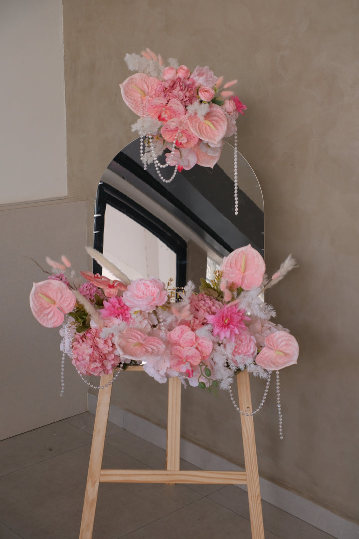 Millen Acrylic Selfie Mirror featuring blush pink artificial florals with ribbon and crystal pearl accents, set on a sleek wooden rack. Customizable display perfect for elegant business events and gifting.