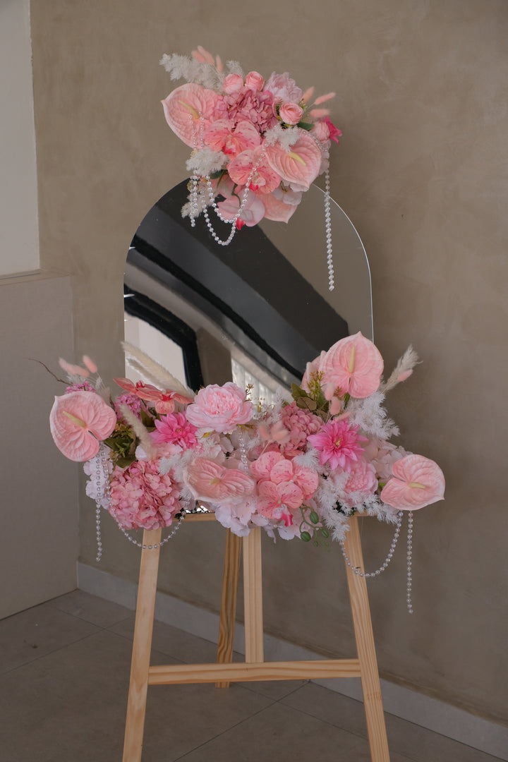 Millen Acrylic Selfie Mirror featuring blush pink artificial florals with ribbon and crystal pearl accents, set on a sleek wooden rack. Customizable display perfect for elegant business events and gifting.