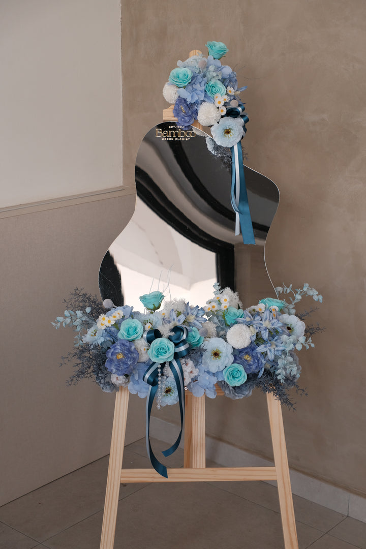 Ophelia Acrylic Mirror featuring a handcrafted blue-themed artificial floral arrangement with elegant ribbons, customizable message panel, and wooden display rack. Perfect for events, gifting, and tabletop decoration.