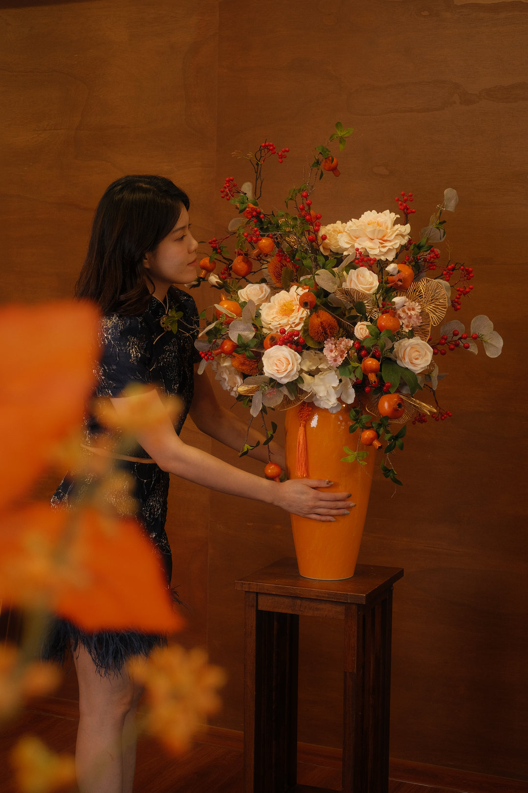Elegant tall orange vase arrangement with artificial cream peonies, faux persimmon branches, and red berries held by a woman in a cheongsam.