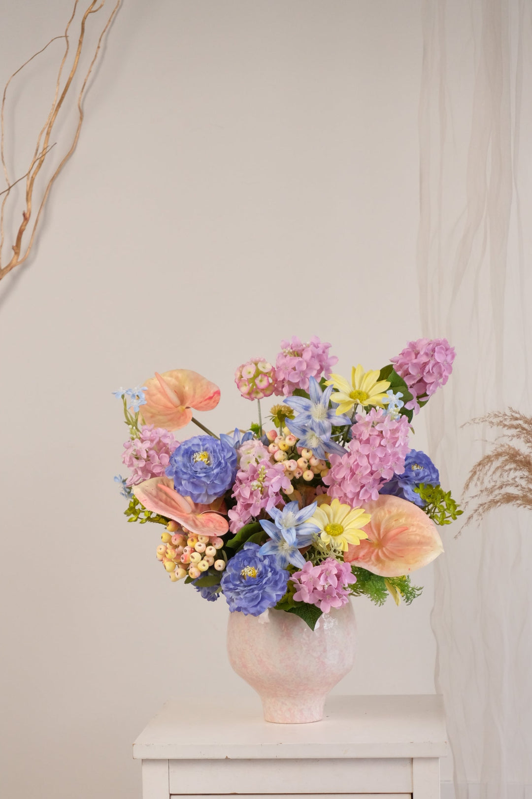 A colorful floral arrangement in a pale pink vase, displayed on a white side table in a styled interior setting with a wood stump, dried grasses, and a neutral wall.

